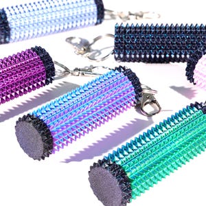 May include: A collection of colorful, spiked keychains in shades of blue, purple, and green. Each keychain features a cylindrical design with a textured, spiky surface and a contrasting dark end cap. The keychains are displayed on a white background.