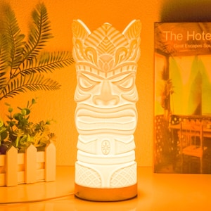 May include: A cream-coloured tiki-style lamp with a detailed carved face and crown. The lamp has a wooden base and is illuminated, casting a warm glow. The lamp is a decorative item.