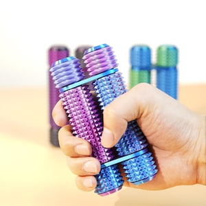 May include: A hand holding two metallic fidget toys. The toys are cylindrical with a textured, spiky surface. One is purple and blue, the other is blue. Other fidget toys in purple, gray, green, and blue are in the background.