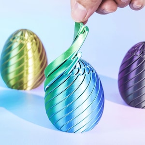 Può includere: Tre sculture metalliche a forma di uovo nei toni del verde, blu e viola. La scultura centrale ha un design a spirale aperto, con una mano che solleva una parte della struttura. Le altre due uova sono piene con un motivo a strisce.