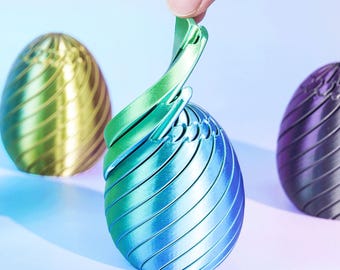 Könnte beinhalten: Drei eiförmige Objekte mit Spiralmuster. Das mittlere Objekt wird angehoben und zeigt ein grün-blaues Farbverlauf-Finish. Links befindet sich ein goldfarbenes Ei und rechts ein dunkelviolettes Ei. Jeder Gegenstand weist eine texturierte Oberfläche auf.