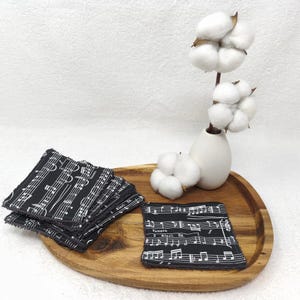 Wiederverwendbare Abschminkpads aus Baumwolle. Obere Seite glatte Baumwolle mit Noten Muster und Rückseite mit Frottee in grau. Liegend auf einem Holzbrett, die Vorderseiten im Noten Muster sind zu sehen, dabei steht eine kleine Vase mit Baumwollpflanzen.