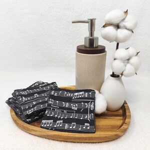 Wiederverwendbare Abschminkpads aus Baumwolle. Obere Seite glatte Baumwolle mit Noten Muster und Rückseite mit Frottee in grau. Liegend auf einem Holzbrett, dabei steht ein Seifenspender und eine kleine Vase mit Baumwollpflanzen.