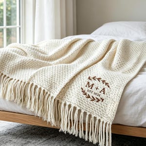 Könnte beinhalten: Eine cremefarbene, texturierte Strickdecke mit langen Fransen an den Rändern. Die Decke zeigt ein braunes Stickmotiv, bestehend aus einem Lorbeerkranz, der die Buchstaben M + A und den Text EST 2026 in der Mitte umschließt.