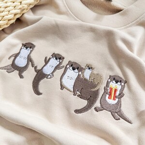 May include: Beige sweatshirt featuring an embroidered design of five otters in various poses. The otters are brown and white, with one holding a gift. The sweatshirt is made of a soft material.