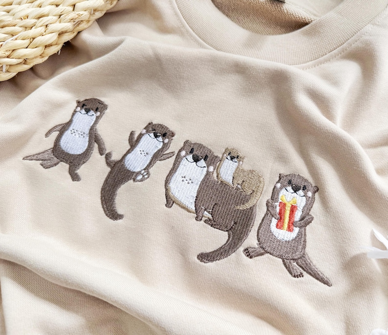 May include: Beige sweatshirt featuring an embroidered design of five otters in various poses. The otters are brown and white, with one holding a gift. The sweatshirt is made of a soft material.