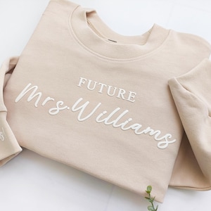 May include: A beige sweatshirt with the text "FUTURE Mrs. Williams" embroidered in white on the front. The cuff of the sleeve has the text "and yes!" embroidered in white.