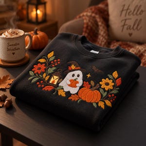 May include: A black sweatshirt featuring an embroidered design of a white ghost wearing a hat, holding a pumpkin, and standing next to a lantern. The graphic includes orange pumpkins, yellow and red autumn leaves, berries, and a small bat.