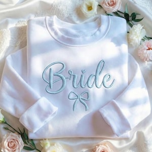 May include: A white sweatshirt featuring the word "Bride" embroidered in a light blue script font, with a bow detail beneath. The garment is presented on a white satin background, decorated with flowers, suitable for a wedding.