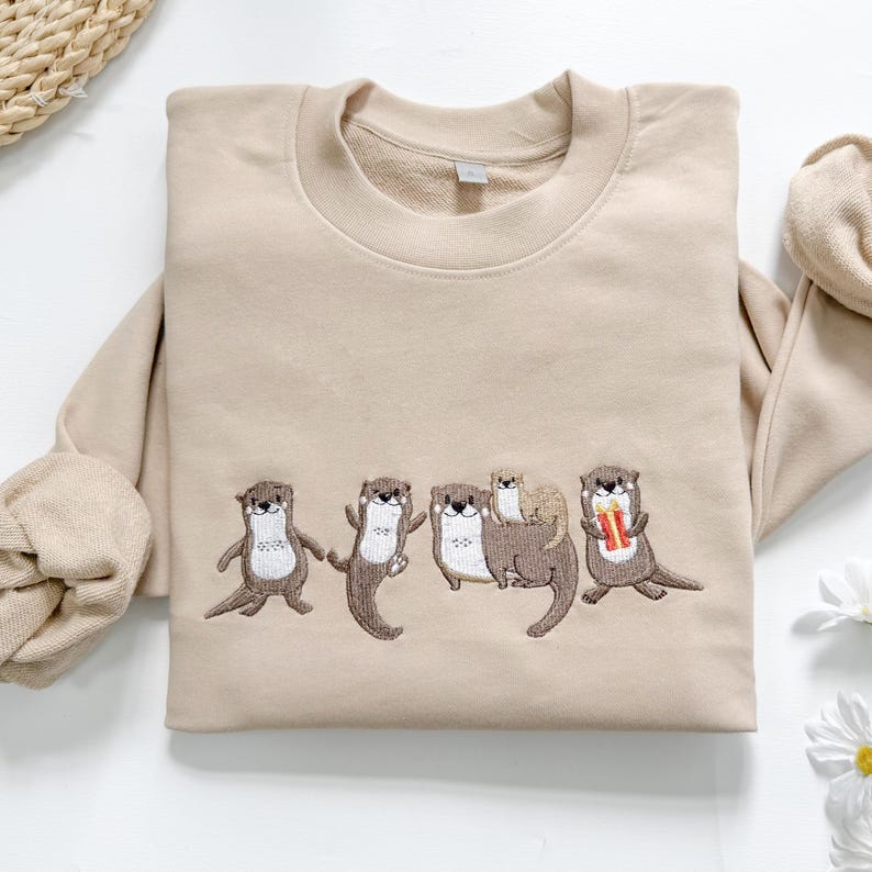 May include: Beige sweatshirt with an embroidered design of five otters. The otters are in various poses, with one holding a gift. The sweatshirt has a crew neck and long sleeves. The fabric is a soft, neutral color.