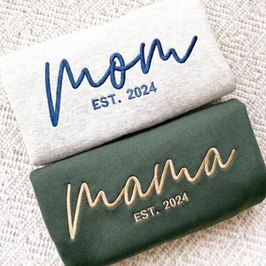 Könnte beinhalten: Zwei Sweatshirts, eines hellgrau und eines dunkelgrün, mit den Wörtern "Mom" und "Mama" in Schreibschrift bestickt. Das hellgraue Sweatshirt hat blaue Stickereien, das dunkelgrüne hat beige Stickereien. Beide sagen "EST. 2024".