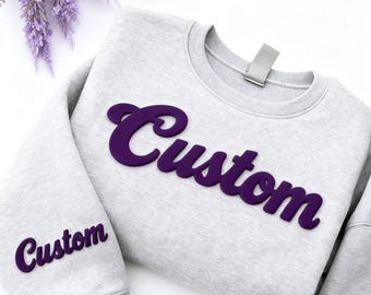 May include: A light gray sweatshirt with the word "Custom" in purple script across the chest and on the left sleeve. The sweatshirt is displayed on a white surface with purple lavender sprigs.
