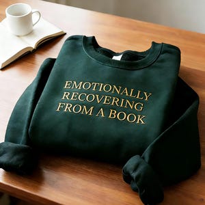 Może przedstawiać: Ciemnozielona bluza z długim rękawem i okrągłym dekoltem, z wyhaftowanym złotym napisem "EMOTIONALLY RECOVERING FROM A BOOK" na środku klatki piersiowej. Odzież posiada prążkowany dekolt, mankiety i d&oacute;ł oraz miękką wyści&oacute;łkę wewnętrzną.
