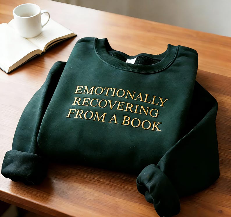 Może przedstawiać: Ciemnozielona bluza z długim rękawem i okrągłym dekoltem, z wyhaftowanym złotym napisem "EMOTIONALLY RECOVERING FROM A BOOK" na środku klatki piersiowej. Odzież posiada prążkowany dekolt, mankiety i d&oacute;ł oraz miękką wyści&oacute;łkę wewnętrzną.