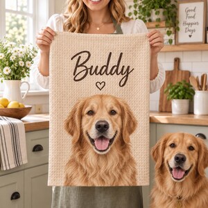 May include: A beige waffle-textured kitchen towel featuring a dark brown printed graphic of a golden retriever dog's face, the name "Buddy" in cursive script, and a small heart icon centred above the dog's head on the fabric surface.