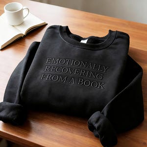 May include: A black sweatshirt with the embroidered text "EMOTIONALLY RECOVERING FROM A BOOK". The garment is folded on a wooden surface, with an open book and a white mug in the background.