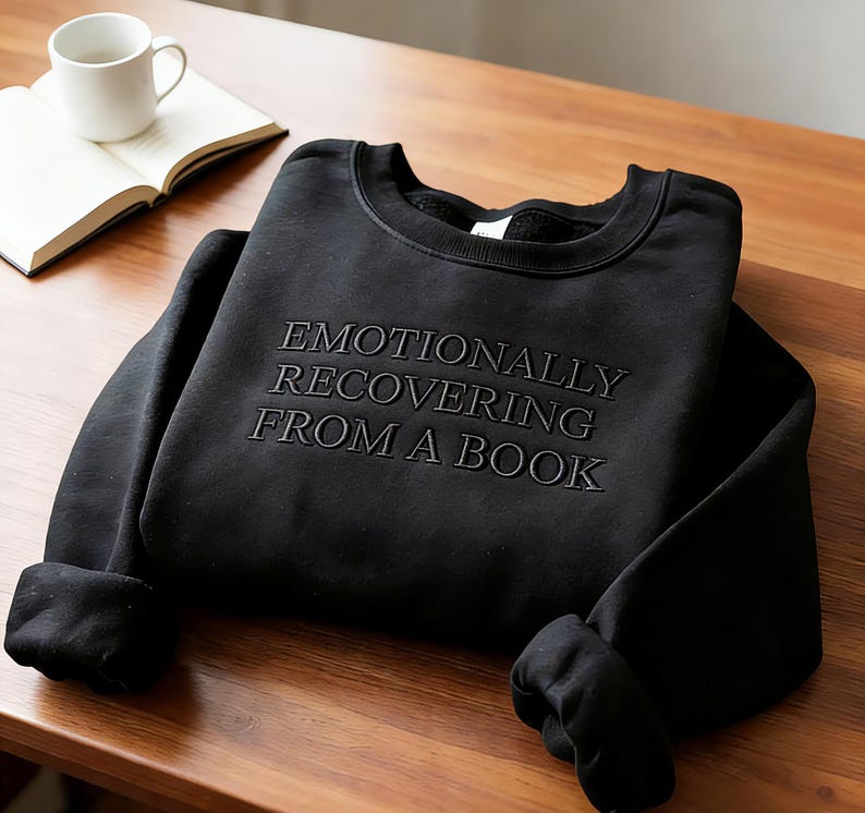 Może przedstawiać: Czarna bluza z okrągłym dekoltem i wyhaftowanym napisem EMOTIONALLY RECOVERING FROM A BOOK pasującą czarną nicią. Odzież posiada długie rękawy z prążkowanymi mankietami oraz prążkowany dekolt, zaprezentowana w złożonej, płaskiej formie.