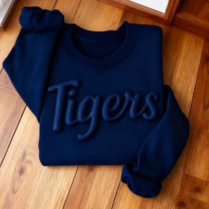 Peut inclure: Un sweat-shirt bleu marine avec le mot "Tigers" en lettres en relief. Le sweat-shirt est plié proprement sur une surface en bois. Les manches sont longues et à poignets. L'encolure ras du cou est également bleu marine.