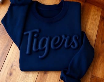 May include: A navy blue sweatshirt with the word "Tigers" in raised lettering. The sweatshirt is folded neatly on a wooden surface. The sleeves are long and cuffed. The crew neck is also navy blue.