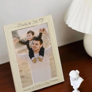 May include: A light wood picture frame with a photo of a father and son. The frame has the text "Daddy & Me" and a heart symbol engraved on the top. The bottom of the frame has the text "Happy 1st Father's Day Daddy".