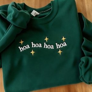 Puede incluir: Una sudadera verde oscuro con la inscripción bordada "hoa hoa hoa hoa" en blanco, acompañada de estrellas doradas. La sudadera está doblada, mostrando la suave tela y el diseño.