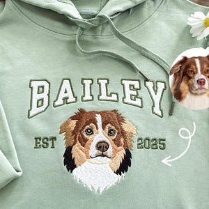 Può includere: Una felpa con cappuccio verde chiaro con il nome "BAILEY" ricamato in bianco e verde. Sotto il nome c'è un ritratto di cane ricamato e il testo "EST 2025". Un piccolo ritratto di cane è nell'angolo in alto a destra.