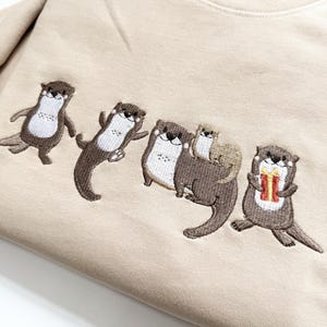 May include: Beige sweatshirt featuring an embroidered design of five otters. The otters are in various poses, with one holding a gift. The embroidery uses brown, white, and red thread.