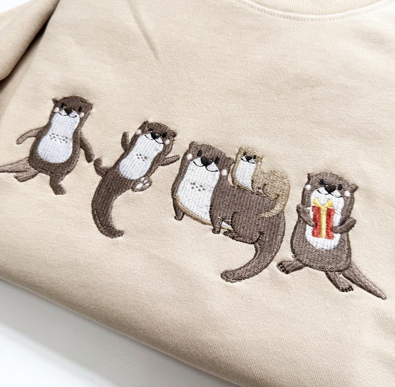 May include: Beige sweatshirt featuring an embroidered design of five otters. The otters are in various poses, with one holding a gift. The embroidery uses brown, white, and red thread.