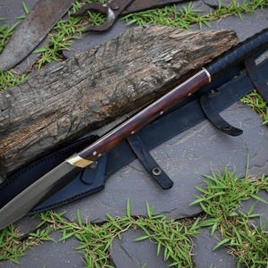 May include: A handmade knife with a dark wooden handle and a sharp, silver blade. The knife rests on a black leather sheath, with a wooden block and other tools in the background. The knife has brass accents.