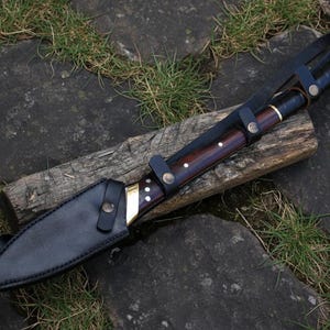 May include: A large, handcrafted knife with a dark wooden handle and a black leather sheath. The blade is partially visible, with a brass bolster and decorative rivets. The knife rests on a piece of wood, with grass and stone in the background.