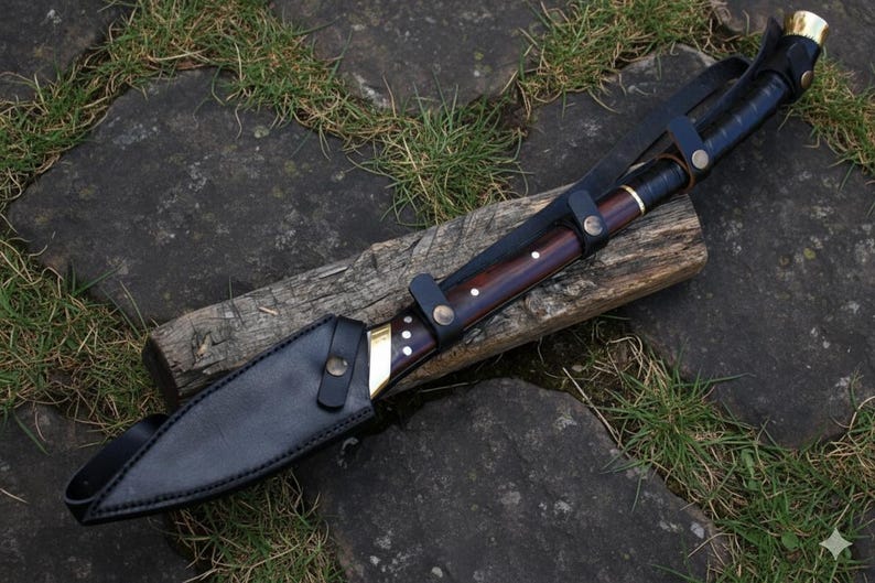May include: A large, handcrafted knife with a dark wooden handle and a black leather sheath. The blade is partially visible, with a brass bolster and decorative rivets. The knife rests on a piece of wood, with grass and stone in the background.