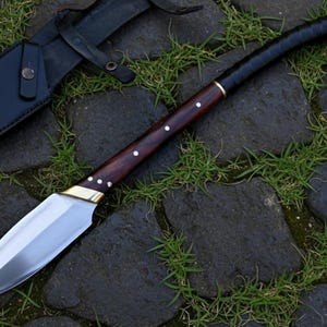 May include: A handmade knife with a polished steel blade and a dark wood handle. The handle is accented with brass and white dots. A black leather sheath is visible in the background. The knife is resting on a stone surface with grass.