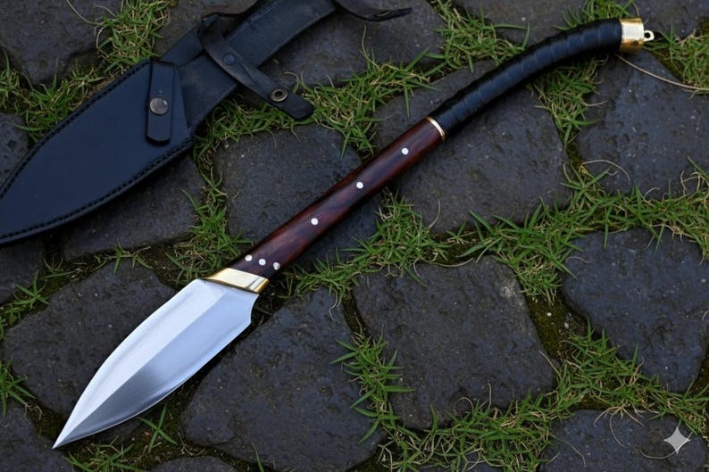 May include: A handmade knife with a polished steel blade and a dark wood handle. The handle is accented with brass and white dots. A black leather sheath is visible in the background. The knife is resting on a stone surface with grass.