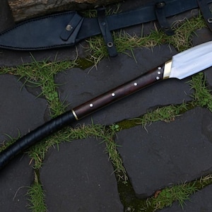 May include: A handcrafted spear with a polished steel blade and a dark wooden shaft. The shaft features brass accents and a black leather-wrapped grip. A black leather sheath is visible in the background.
