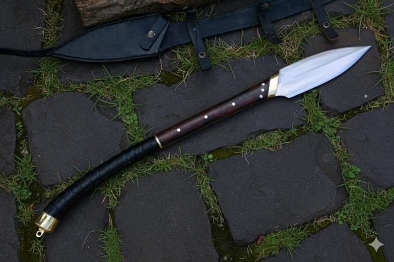 May include: A handcrafted spear with a polished steel blade and a dark wooden shaft. The shaft features brass accents and a black leather-wrapped grip. A black leather sheath is visible in the background.