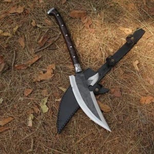 May include: A large, handmade knife with a curved blade and a dark brown handle. The knife is in a black leather sheath, resting on a bed of dry leaves and pine needles. The blade has a silver finish.