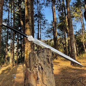 Pode incluir: Uma espada longa e prateada com uma empunhadura preta repousa sobre um tronco de árvore. A lâmina é altamente polida, refletindo a floresta circundante. O design da espada apresenta uma guarda e uma ponta, sugerindo um tema medieval ou de fantasia.