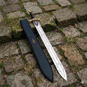 May include: A silver sword with a decorative blade and a brown and gold handle lies next to its black leather sheath. The sheath and the sword's hilt feature a gold emblem. The sword is displayed on a stone surface.