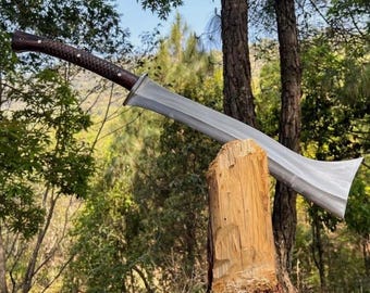 Puede incluir: Una espada grande, de color plateado, con una hoja ancha y un mango marrón oscuro. La espada está apoyada en un tocón de madera en un entorno forestal, con árboles y follaje en el fondo. El mango tiene una apariencia texturizada y tejida.