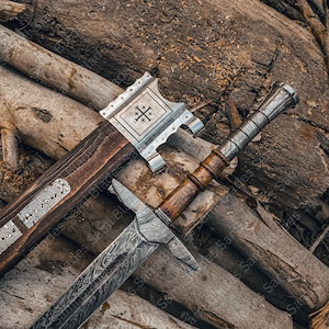 Close up view of the top handle of a hand forged Damascus steel Knight Templar Sword, showing polished hardwood grip, steel fittings, part of the patterned blade, and the upper section of the matching scabbard