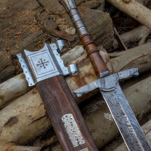 Close up view of the top handle of a hand forged Damascus steel Knight Templar Sword, showing polished hardwood grip, steel fittings, part of the patterned blade, and the upper section of the matching scabbard