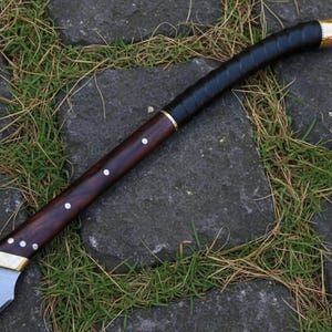 May include: A handcrafted sword with a polished steel blade and a dark wood handle. The handle is wrapped in black leather and accented with brass fittings. The sword is lying on a stone surface with grass.