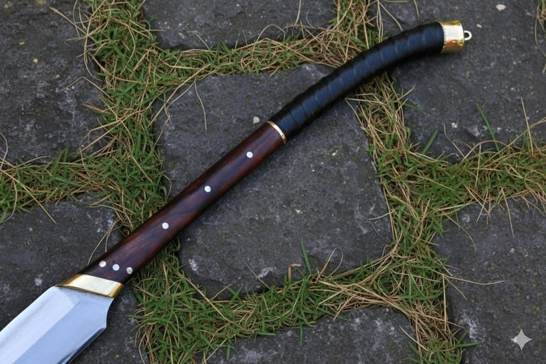 May include: A handcrafted sword with a polished steel blade and a dark wood handle. The handle is wrapped in black leather and accented with brass fittings. The sword is lying on a stone surface with grass.