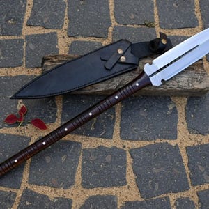 May include: A spear with a polished, double-edged blade and a dark wooden shaft. The shaft is adorned with small, white studs. A black leather sheath rests beside the spear on a wooden plank. The spear is approximately 70 inches long.