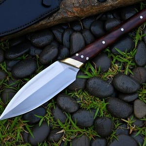 May include: A handmade spear point knife with a polished steel blade and a dark wood handle. The handle is accented with brass and white rivets. The knife rests on a bed of black stones and green grass, with a black leather sheath visible.