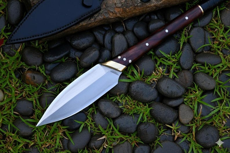 May include: A handmade spear point knife with a polished steel blade and a dark wood handle. The handle is accented with brass and white rivets. The knife rests on a bed of black stones and green grass, with a black leather sheath visible.