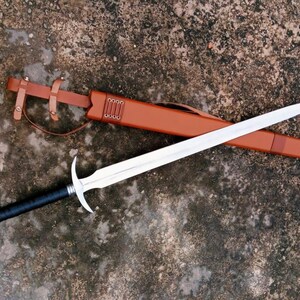 May include: A long, silver sword with a black handle and a silver pommel rests on a brown leather sheath. The sheath is secured with a brown leather strap. The sword is a medieval-style weapon.