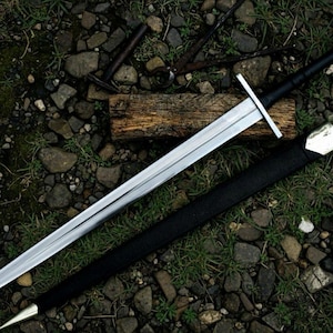 May include: A long, silver sword with a black handle and crossguard, lying on a bed of rocks and grass. The sword's scabbard, also black, is beside it. The blade is highly polished, reflecting light. A piece of wood is behind the sword.