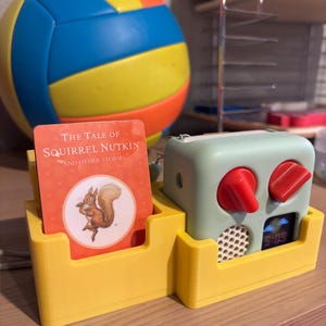 May include: A yellow 3D-printed organizer holds a card with a squirrel illustration and text "The Tale of Squirrel Nutkin". A mint green clock with red knobs and a digital display showing "5:05" sits beside it. A blue, yellow, and orange volleyball is in the background.