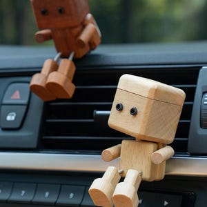May include: Two wooden robot figurines, one light and one dark, designed to sit on a car's air vent. The robots have movable arms and legs, adding a playful touch to the vehicle's interior.